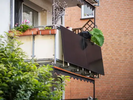Auf dem Balkon eines Mehrfamilienhauses steht ein Mann und winkt; auf dem Dach über ihm sind zahlreiche Photovoltaikmodule installiert.