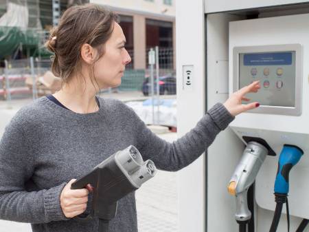 Eine junge Frau mit grauem Pullover steht an einer öffentlichen Ladesäule und tippt auf deren Display.