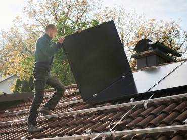 Ein Solateur auf einem Dach trägt ein PV-Modul, darauf sind Monatgeschienen, auf denen bereits viele Module installiert sind