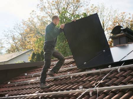Ein Solateur auf einem Dach trägt ein PV-Modul, darauf sind Monatgeschienen, auf denen bereits viele Module installiert sind