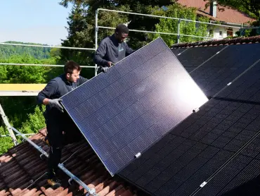 Zwei Personen montieren auf einem Schrägdach ein Modul einer PV-Anlage, dahinter ein Baugerüst, ein weiteres Haus und bewaldete Hügel.
