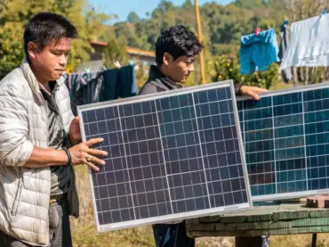 Zwei junge Männer mit Solarpanelen an einem Tisch im Freien; im Hintergrund trocknende Wäsche auf einer Leine.