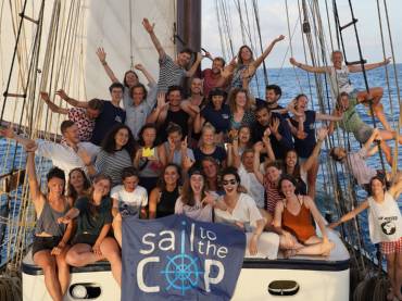 Eine Gruppe gutgelaunter junger Menschen auf dem Deck eines Segelschiffs, vor ihnen ein Banner mit der Aufschrift «Sail to the COP»