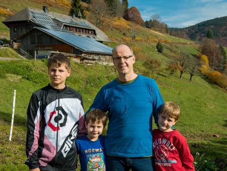 Ein Vater mit drei Söhnen steht vor einem Hang, im Hintergrund ist eine Scheune mit Solardach zu sehen