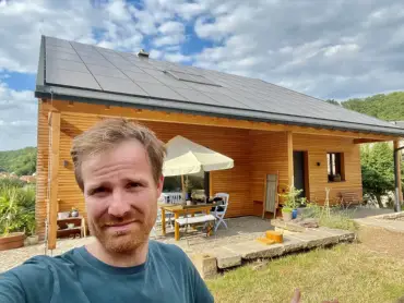 Selfie: Ein junger Mann vor einem freistehenden Holzhaus; das Dach ist vollständig mit PV-Modulen belegt.