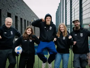 Fünf junge Menschen in Vereinskleidung stehen bei einem Stadion und lachen in die Kamera.