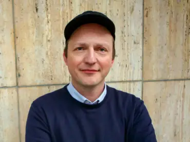Ein Mann mit Schirmmütze und blauem Pullover vor einer Wand aus Kalkstein.
