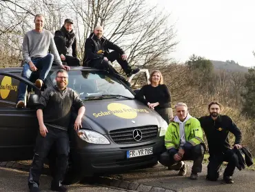 Ein Team aus sieben Personen um und auf einem kleinen schwarzen Lieferwagen am Wegesrand, auf dem Lieferwagen das Logo «Solarbau Freiburg».