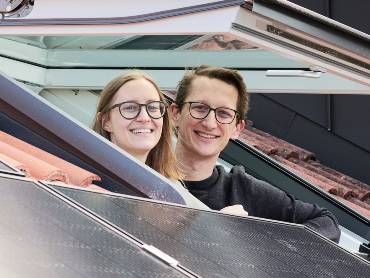 Ein junges Pärche schaut gut gelaunt und stolz aus einem Dachfenster, neben dem PV-Module zu sehen sind. 