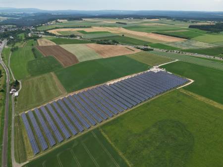 Blick auf ein Solarkraftwerk mit über 30 Modulreihen, im Hintergrund ein Dorf inmitten flach gewellter Landschaft