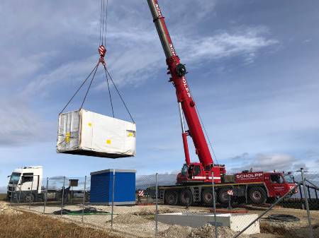 An einem Schwerlastkran hängt ein weißer Container, der die Trafostation für den Solarpark enthält