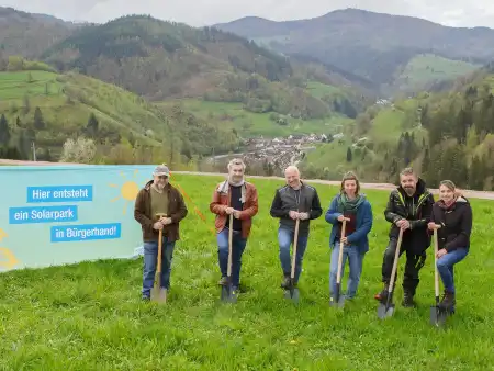 Neben einem Plakat, das über den Bau des Solarparks informiert, stehen sechs Personen mit Spaten auf einer steilen Wiese; hinter ihnen eine malerische Schwarzwaldkulisse