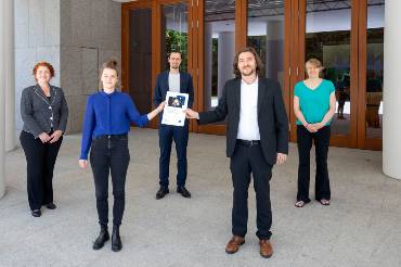 Mit coronabedingtem weitem Abstand stehen fünf Menschen zum Gruppenfoto vor einem Gebäudeeingang aufgestellt, zwei halten gemeinsam ein Dokument hoch