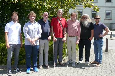 Gruppenfoto von sieben Männern nebeneinander auf der Straße vor einer Hecke