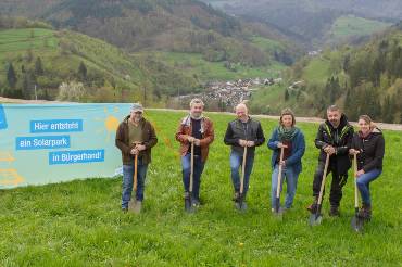 Gruppe von sechs Menschen mit Spaten nebeneinander neben einem Banner