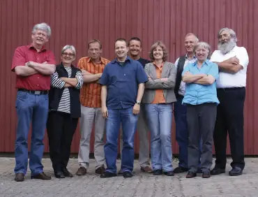 Ein gutes Dutzend Menschen steht, locker und freundlich zum Foto gruppiert, vor einer braunrot gestrichenen Holzwand.