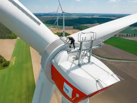 Ein Monteur steht in luftiger Höhe auf einer EWS-Windkraftanlage, zwischen den Feldern dahinter stehen zwei weitere Anlagen.