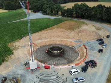 Luftbild: Windenergieanlage im Bau; die Stahlarmierungen des kreisförmigen Fundaments sind bereits montiert, darauf tummeln sich einige Bauarbeiter, daneben steht ein Kran.