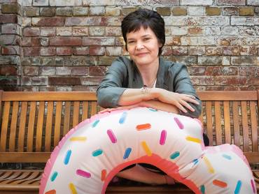 Auf einer altmodischen Holzbank vor einer rustikalen Ziegelmauer sitzend, verschränkt die lächelnde Kate Raworth ihre Arme in Brusthöhe auf einem Schwimmreifen mit Donut-Dekor, der vor ihr auf dem Boden steht.