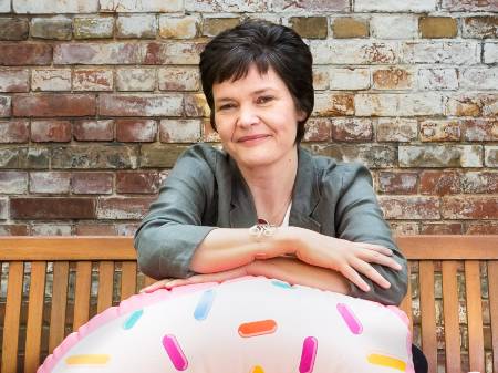 Auf einer altmodischen Holzbank vor einer rustikalen Ziegelmauer sitzend, verschränkt die lächelnde Kate Raworth ihre Arme in Brusthöhe auf einem Schwimmreifen mit Donut-Dekor, der vor ihr auf dem Boden steht.