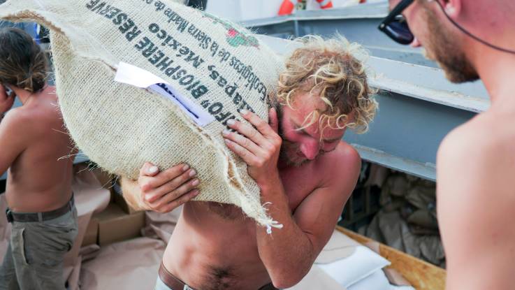 Ein Mann mit nacktem Oberkörper und vor Anstrengung gerötetem Gesicht schleppt einen Sack mit Waren, auf dem die Worte «Biosphere Reserve» zu lesen sind.