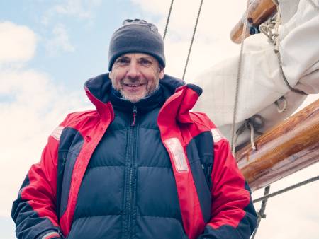 Unter dem Großbaum eines Segelschiffes lächelt ein Mann mit wettergegerbtem Gesicht unter seiner Wollmütze vor, er trägt eine Steppjacke.