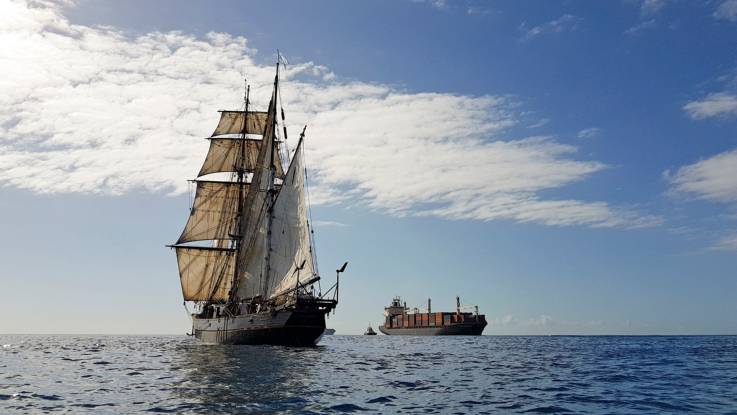 Ein Zweimaster mit klassischer Takelage, von der Bugseite her aufgenommen, begegnet einem Containerfrachter auf offener See.
