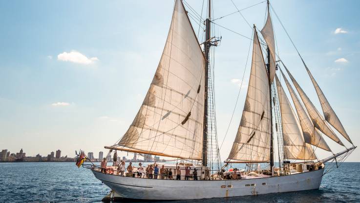 Ein Segelschiff mit deutscher Flagge und unter voller Takelage nimmt Fahrt auf, während die Besatzung winkend an der Reling steht; im Hintergrund eine Stadt mit einigen Hochhäusern.