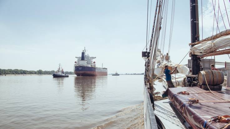 Während an Bord des Segelschiffs die Hauptsegel verzurrt werden, wird ein riesiger Frachter meerwärts geschleppt