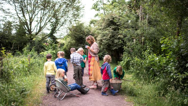 Auf einem Waldweg tummeln sich Kinder, beobachtet von einer Erzieherin. Ein blondes Mädchen sitzt in einer umgedrehten Schubkarre und isst Eis.