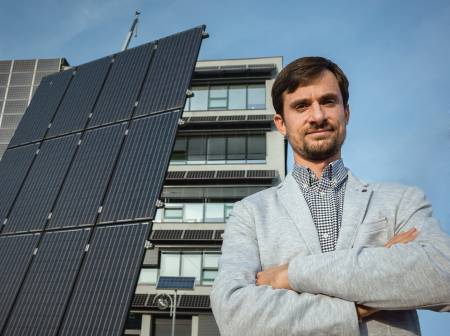 Ein bärtiger junger Mann steht mit verschränkten Armen neben einer PV Anlage und schaut entschlossenen Blickes in die Kamera.