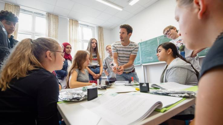Ein junger Mann sitzt auf einem Tisch und erklärt  einer Gruppe von zehn Schülern und Schülerinnen etwas.