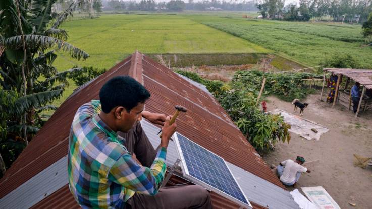 Auf einem Hausdach sitzt ein Mann und installiert ein kleines Solarpanel. Im Hintergrund sind grüne Felder zu sehen.