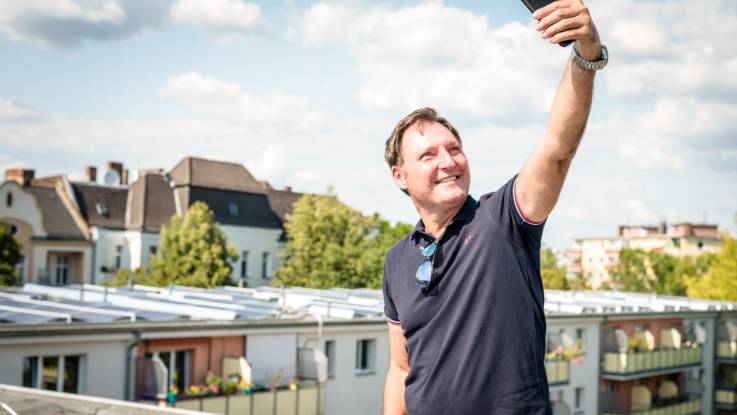 Auf einem Flachdach steht ein Mann mittleren Alters. Er hält mit der linken Hand sein Smartphone hoch, um ein Selfie zu schießen.