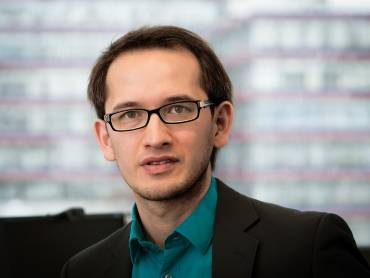 Kopfportrait eines jungen Mannes mit Brille vor städtischer Kulisse.