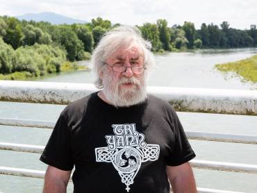 Auf einer Brücke steht ein Mann mit grauem Rauschebart und blinzelt in die Kamera.