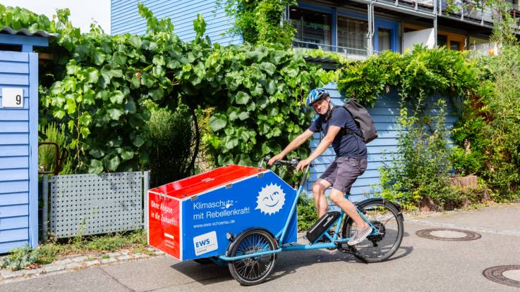 Ein Mann fährt auf einem Lastenfahrrad vor begrünten Häusern entlang.