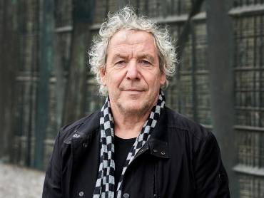 Mann mit zerzausten grauen Haaren steht vor einer Wand und schaut in die Kamera.