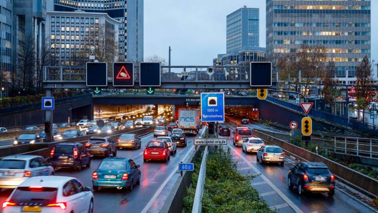 Ein mehrspurige Straße in einer Großstadt im Regen, auf der vor einem Tunnel Auto an Auto im Stau steht.