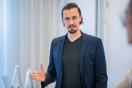 Junger Mann mit Bart im blauen Jacket spricht und gestikuliert entspannt in einer Präsentation.
