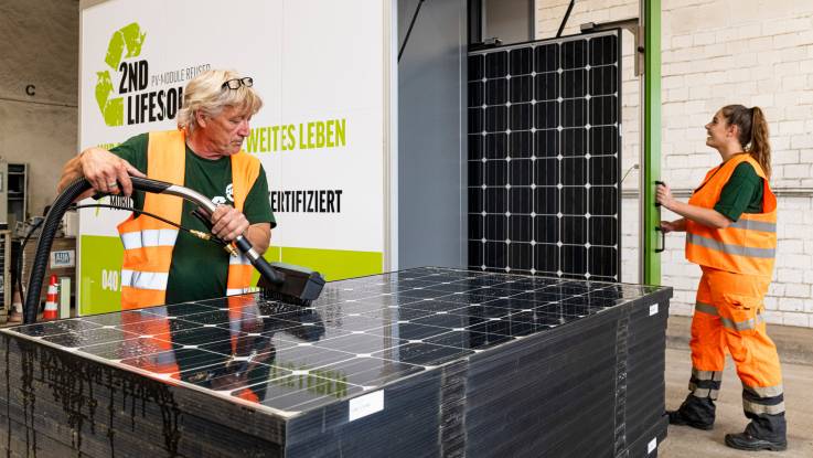 Eine Person reinigt mit einer Wasserbürste ein auf einem Stapel liegendes PV-Modul. Im Hintergrund zieht eine Frau Module aus einem Wagen.