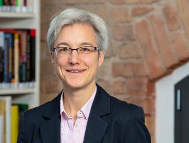 Portrait einer Frau mit kurzen grauen Haaren vor einer Backsteinwand.