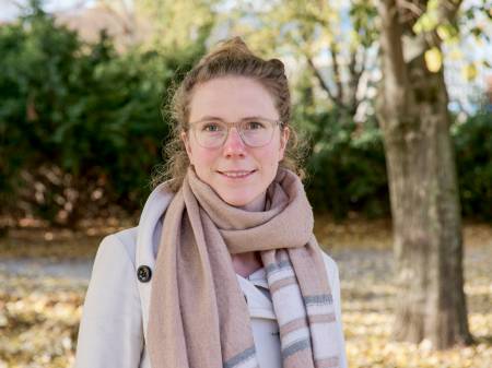 A friendly-looking young woman stands in front of autumnal trees and smiles into the camera.