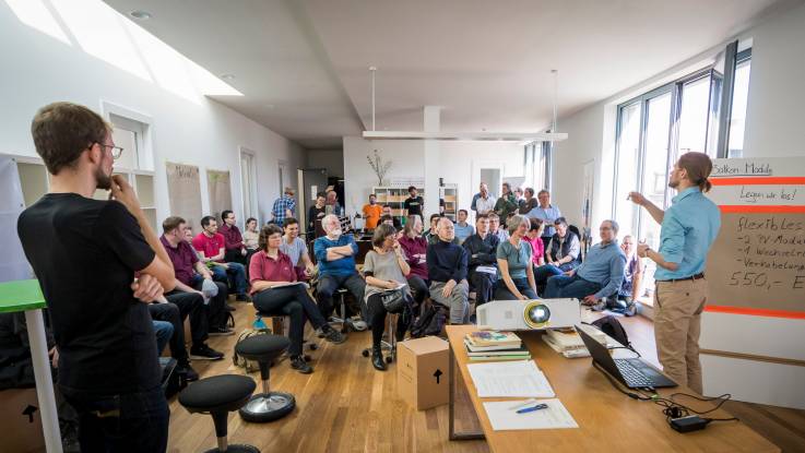 View of a bright spacious room, where some 40 people are listening to a presentation being given by two men in the foreground.