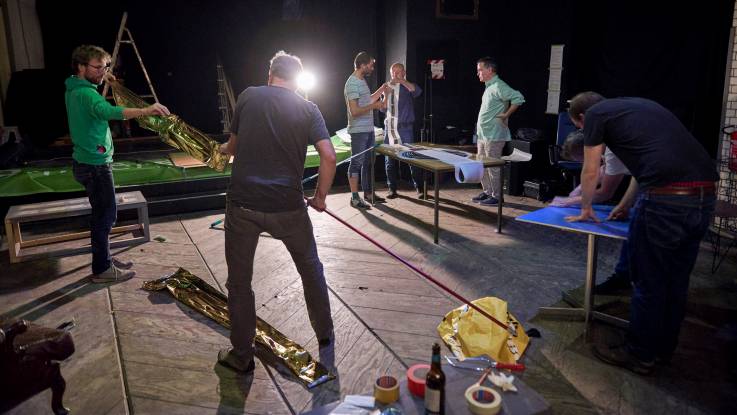 In a sparsely lit large room, six young men work on making paper and cardboard signs.