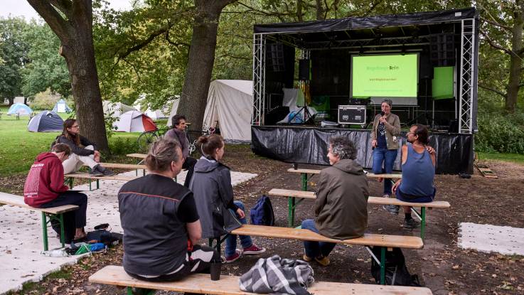 Several spectators sit on fold-up benches beneath tall trees in front of a stage during a discussion.