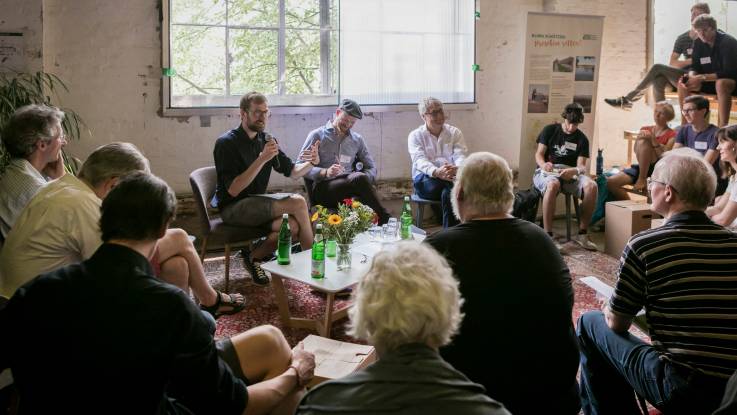 People are sat in a circle having a discussion in a relaxed atmosphere around a small coffee table in a room full of natural light.