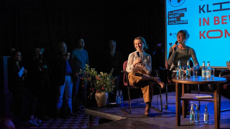 Two young women sit on a blue-lit stage and talk to the audience.