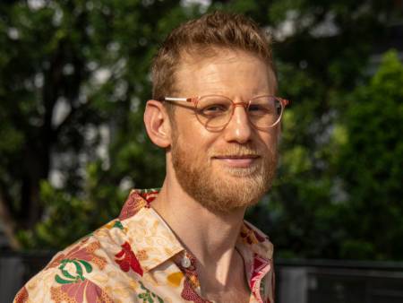 Portrait eines jungen Mannes mit kurzem rotem Haar, gepflegtem Vollbart und runder Brille draußen vor Bäumen. 