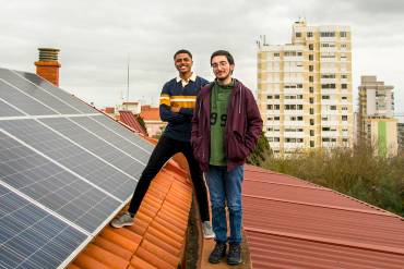 In the midst of a rooftop landscape with PV systems, two young men stand talking happily.